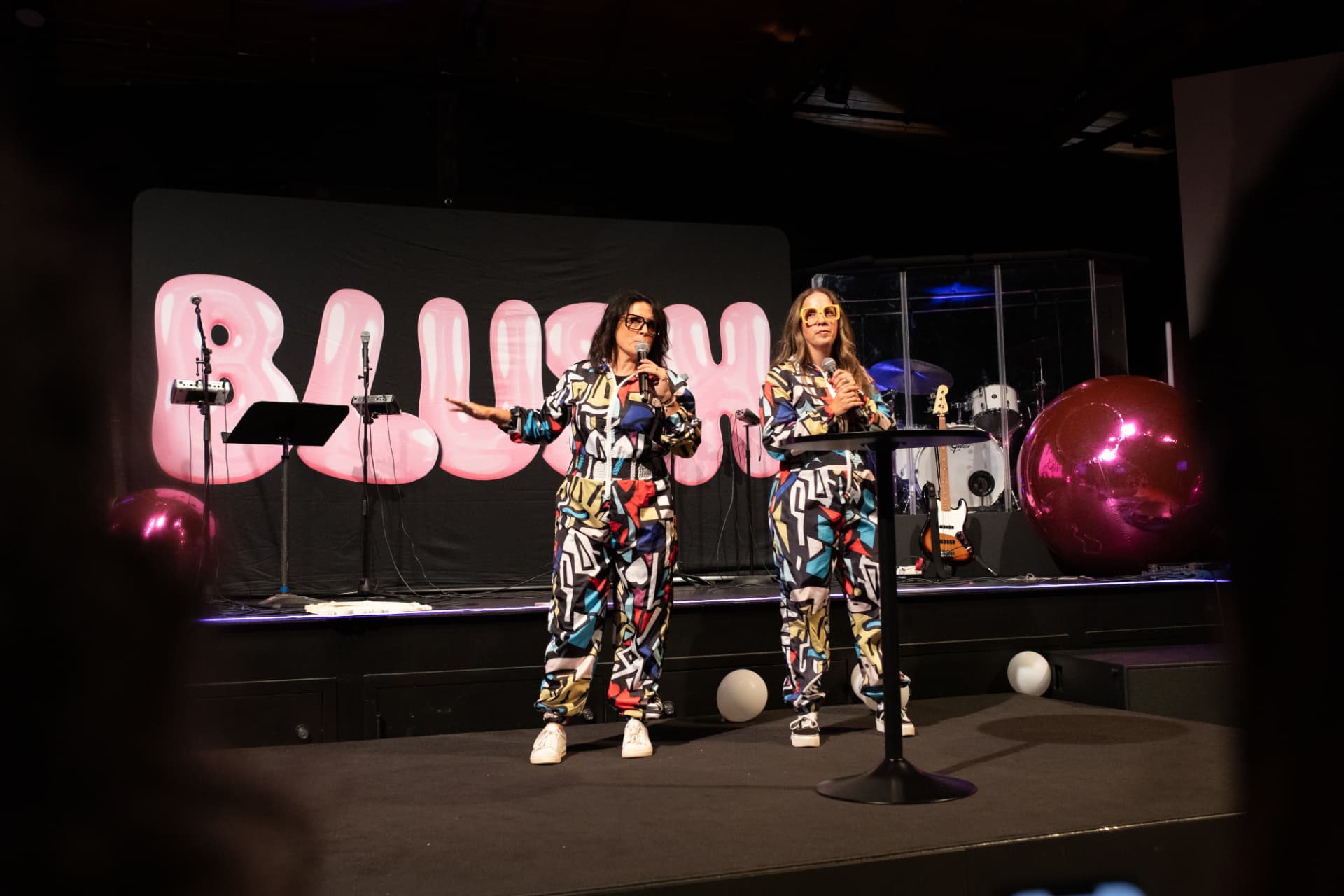 Two BLUSH hosts in colorful outfits on stage with the pink logo backdrop
