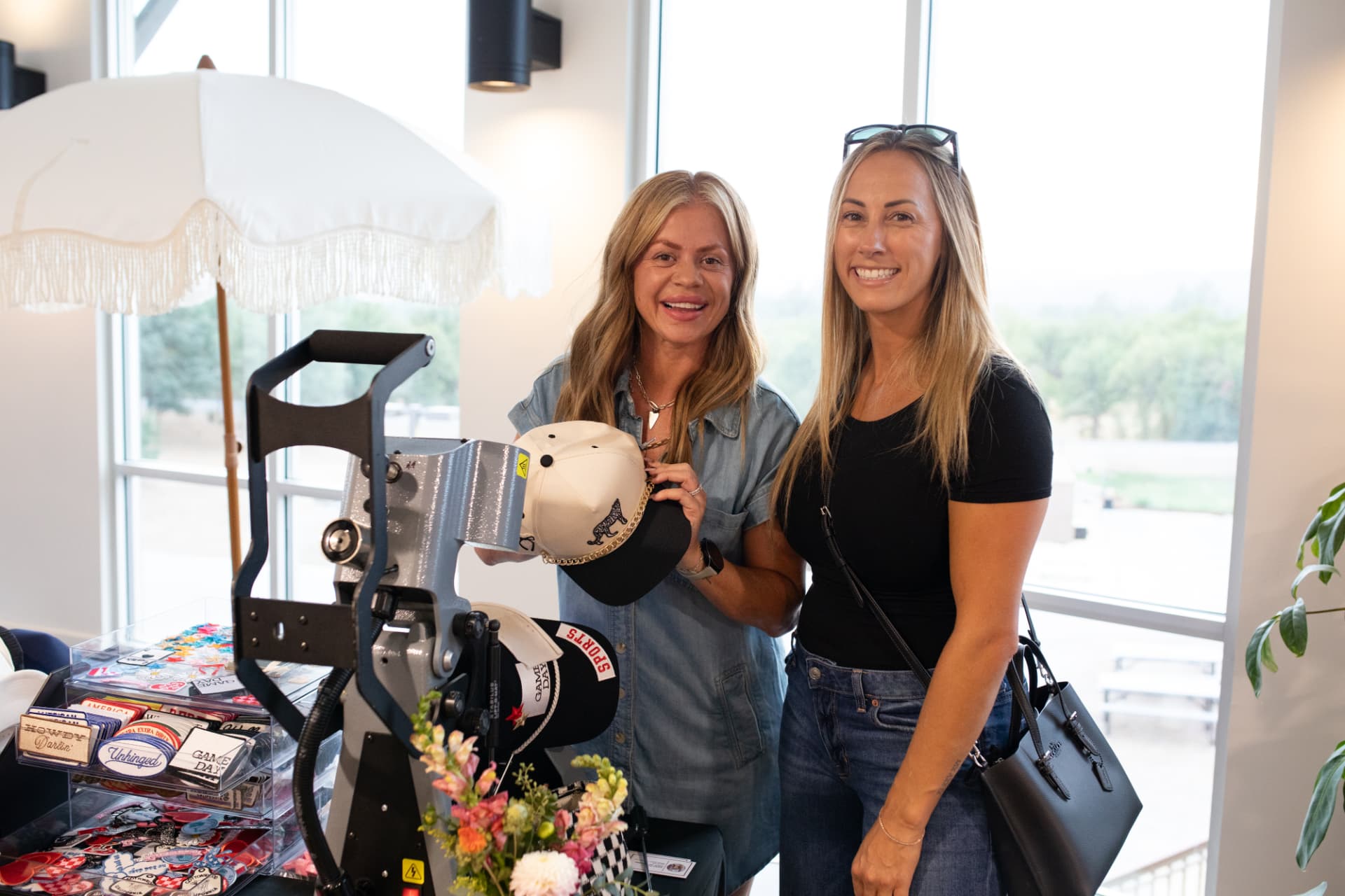 Two women smiling beside a custom hat press at BLUSH